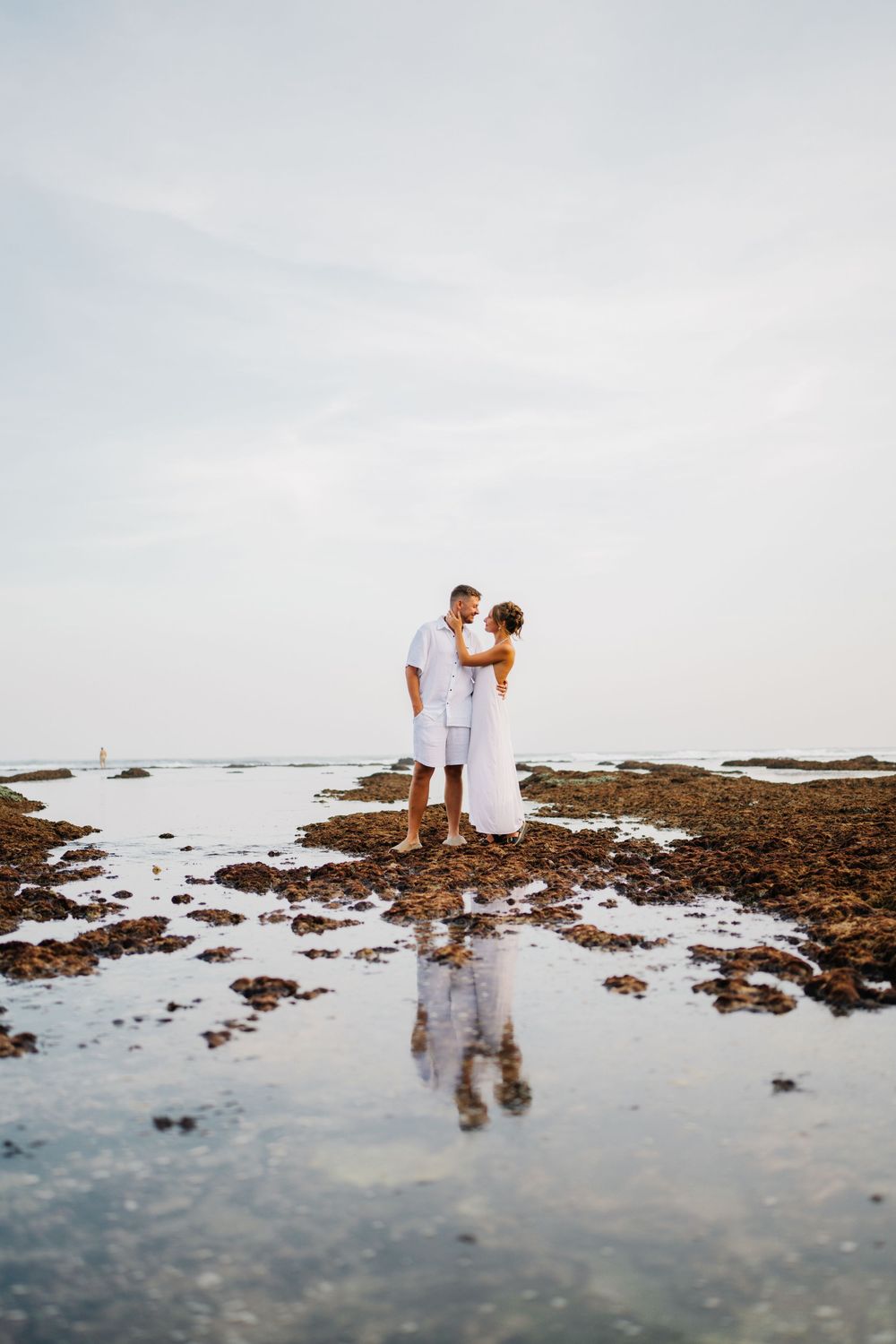 A couple in white stands on rocky tide pools with their reflection in the water.
