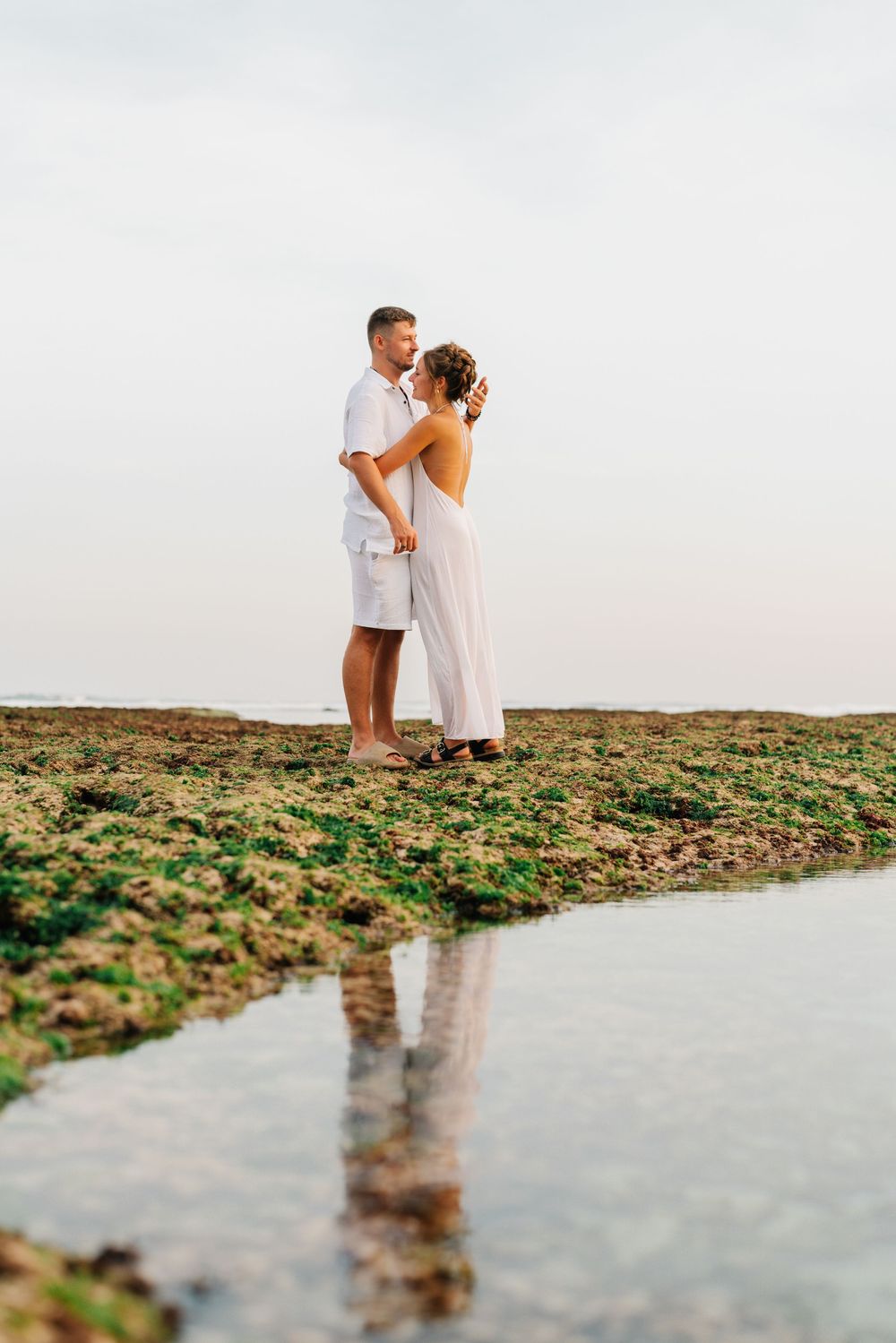 Two people in white outfits share a tender moment by the water's edge.
