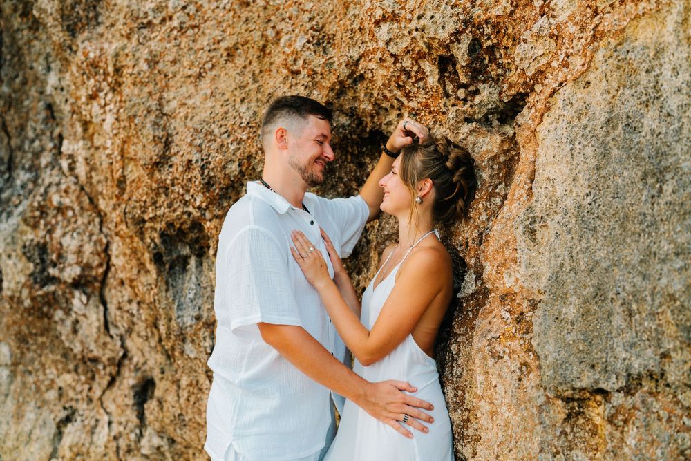 A romantic couple in white outfits embraces against a rocky cliff wall during sunset.