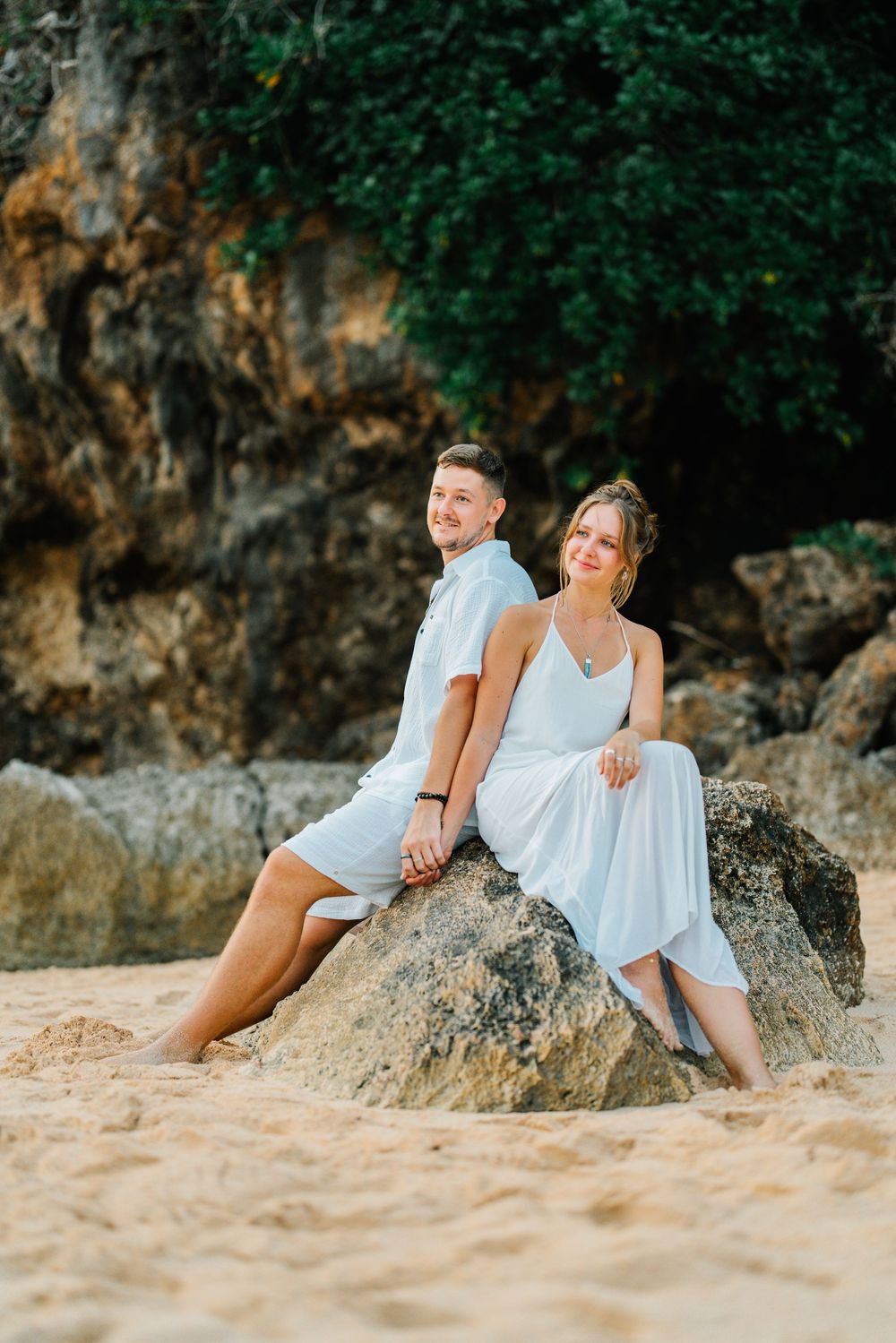 A couple in white outfits relaxing on a sandy beach against rocky cliffs.