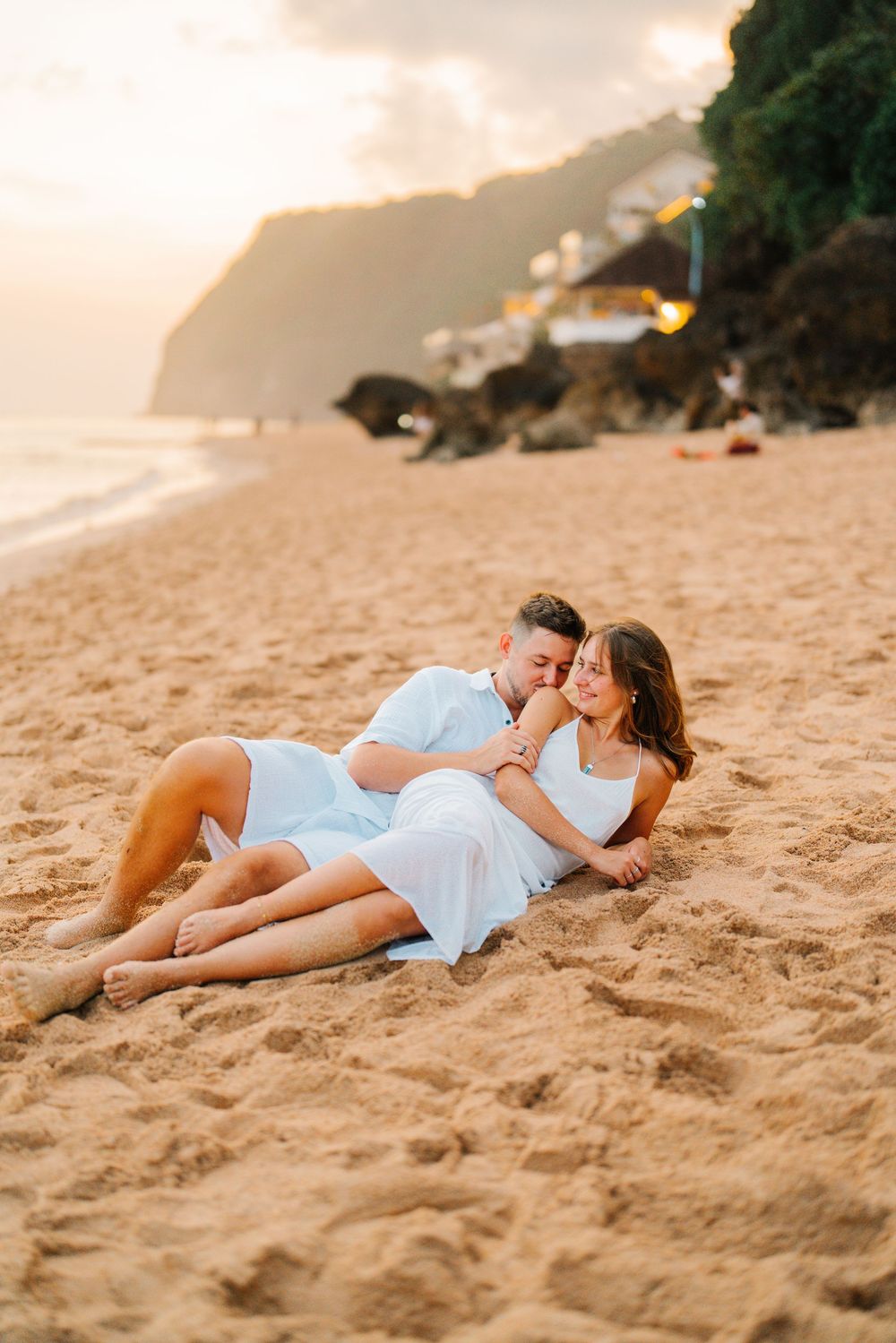 A couple in white clothing lies together on golden sand at the beach during sunset.