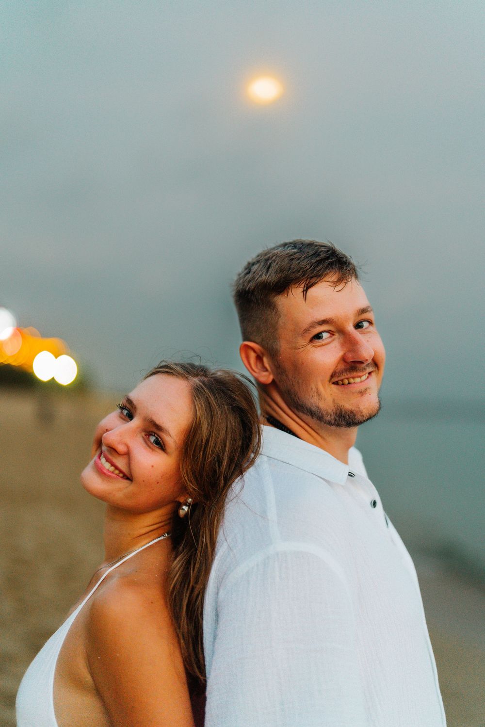 A couple in white outfits shares a tender moment against a dusky beach backdrop.