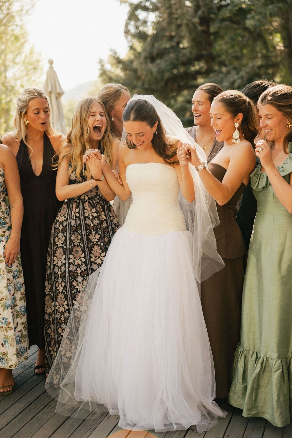 Wedding party laughs together outdoors wearing sage green and black dresses with a bride in white.