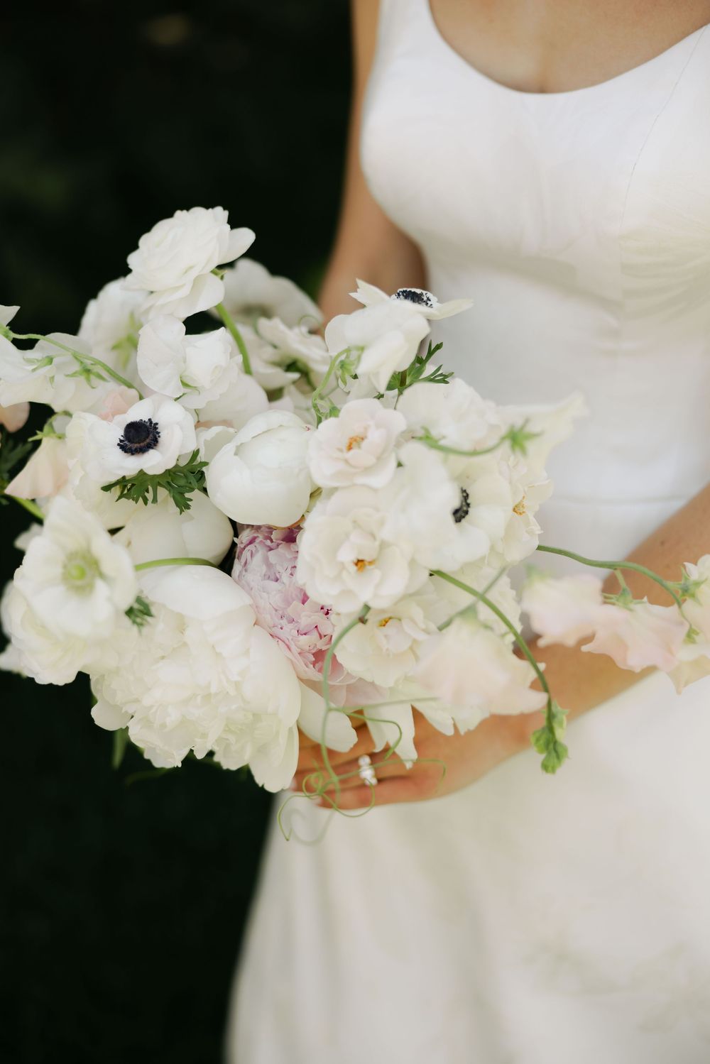 A delicate white and blush wedding bouquet with anemones and roses held against a white dress.