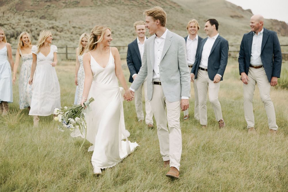 Wedding party in white and beige attire walking through a grassy mountain landscape.