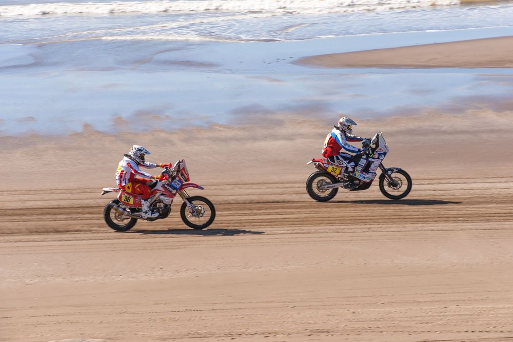 Motorcycles racing across a sandy beach with ocean waves in the background during a competitive event.