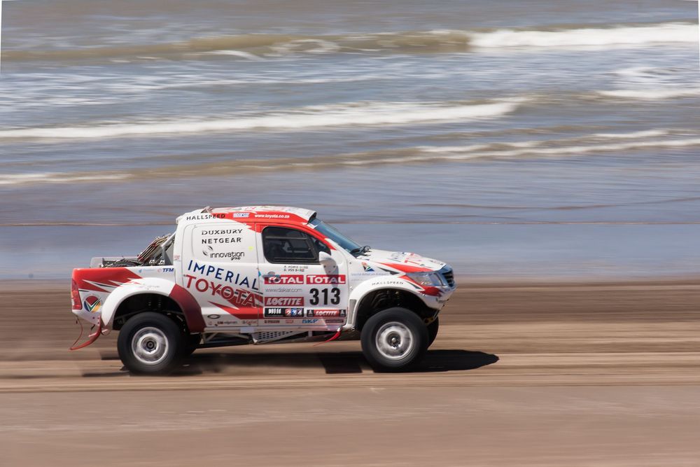 A red and white rally car races across a sandy beach track at high speed leaving a trail of sand in its wake.