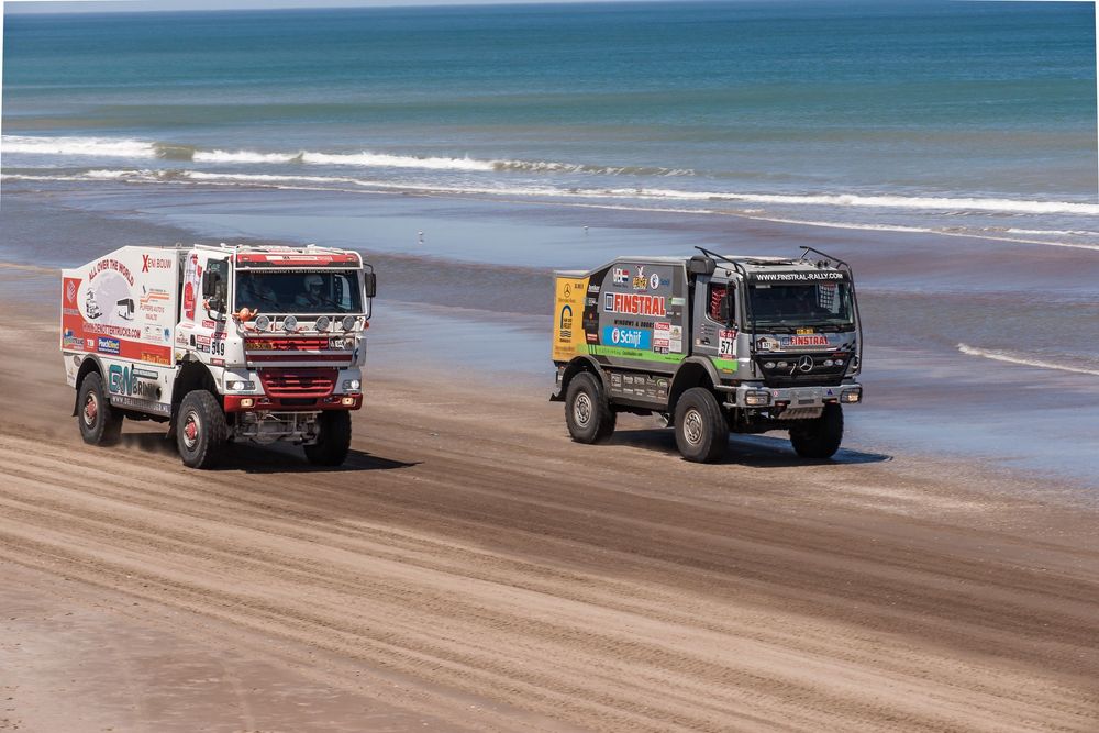 Racing trucks speed across a sandy beach with blue ocean waters visible in the background during a coastal competition.