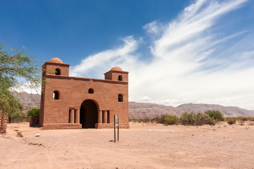 Adobe brick structure with arched entrance stands isolated in arid desert landscape against blue sky and mountains.