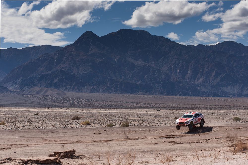 Panoramic desert landscape with rugged mountains, sparse vegetation, and cloud-scattered blue sky stretches into horizon.