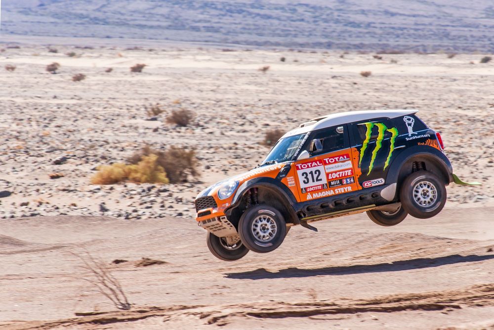 Rally cars and buggy vehicles racing through a dusty desert terrain with rocky mountains in the distance.