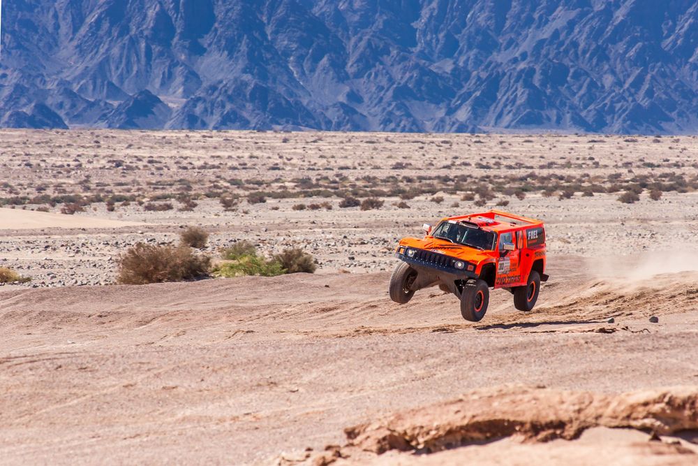 Remote controlled orange rock crawler tackles steep terrain obstacle course in desert mountain landscape.