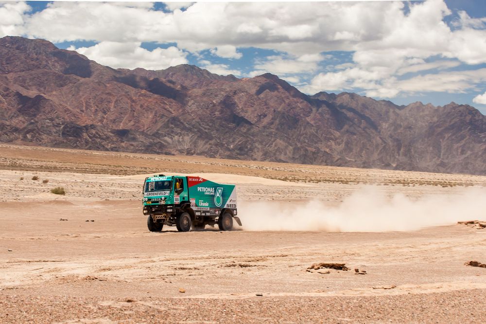 Green supply truck driving through vast desert terrain with mountains and cloudy skies in the background.