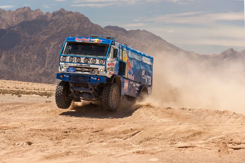 Blue off-road racing truck kicks up dust while racing through desert terrain against mountain backdrop.