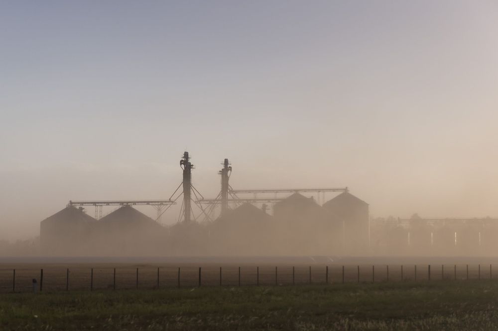 Foggy morning landscape showing silhouettes of farm buildings and barns against a misty rural horizon at sunrise.