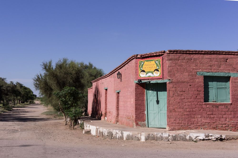 Pink adobe buildings with turquoise doors line a dusty desert road under bright blue skies in the American Southwest.