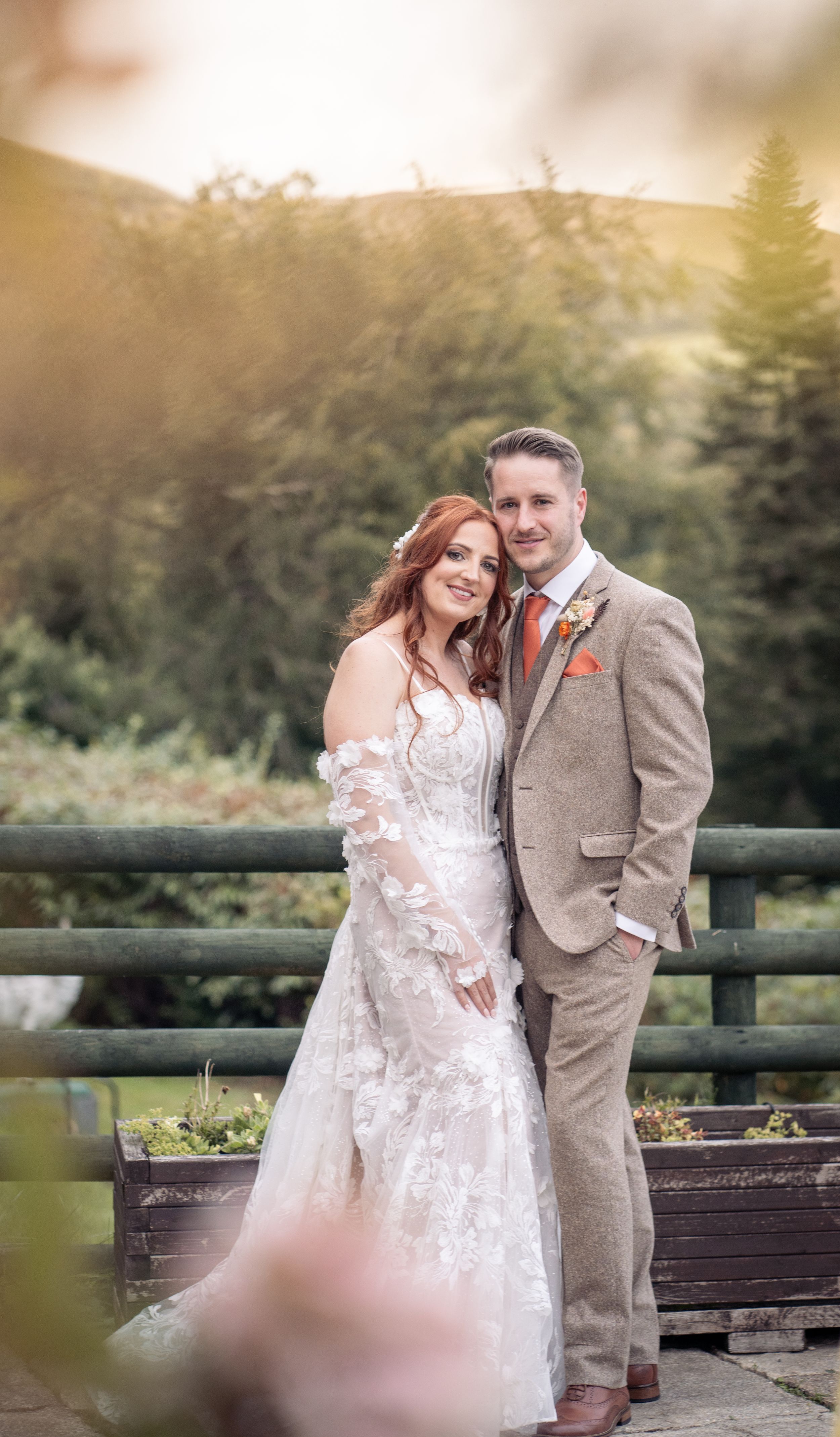 A romantic wedding couple stands together by a wooden fence during sunset in an autumn outdoor setting.