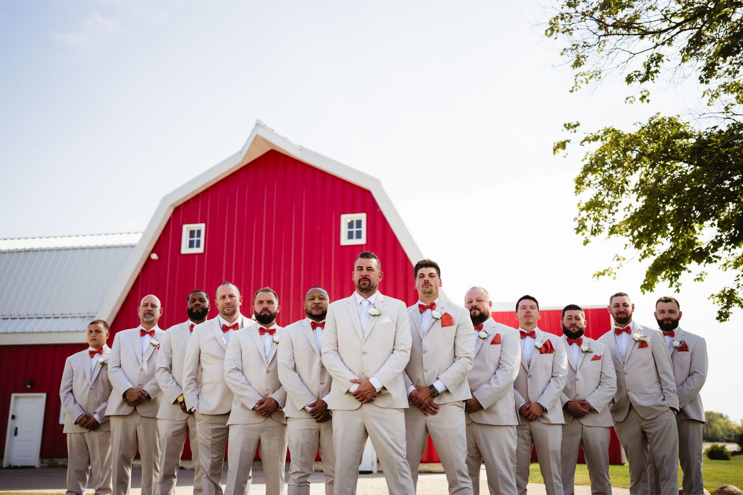A group in light beige suits poses in front of a bright red barn on a sunny day.
