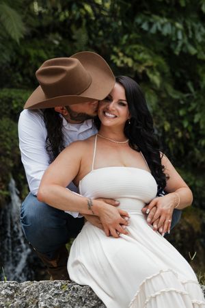 Couple in white outfits pose by waterfall, man in cowboy hat kisses smiling brunette woman.