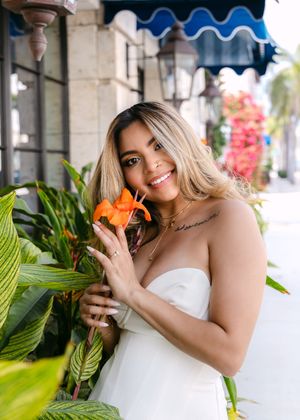 A bride in a white strapless dress poses outdoors against tropical foliage and pink flowers at a venue.