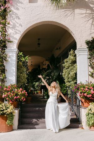 A person in a white dress poses playfully under a series of Spanish-style archways adorned with pink flowers and greenery.
