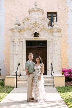 A couple stands in front of an ornate Spanish colonial church entrance with white stone details and pink bougainvillea.