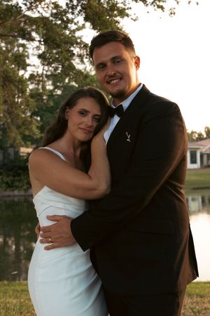 A couple in wedding attire embracing outdoors, man in black tuxedo and woman in white dress, near a pond at sunset.