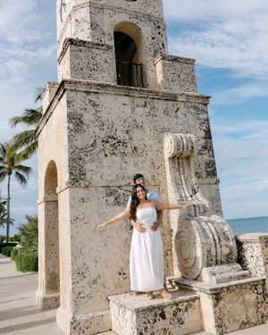 Woman in white dress poses by historic stone tower structure near beach.