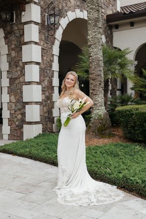 Smiling bride in a white lace wedding gown holding a bouquet of white lilies, posing outdoors near palm trees.