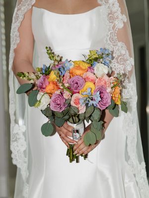 Bride in white satin gown holding colorful wedding bouquet with pink, orange, yellow roses and blue flowers with lace veil.