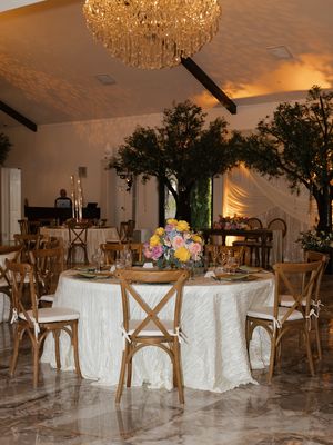 Elegant wedding reception venue with rustic wooden chairs, white linen tables, floral centerpieces, tall trees, and a crystal chandelier.
