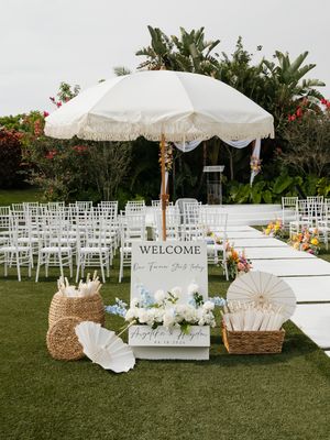 Outdoor wedding ceremony setup with white chairs, floral arrangements, a welcome sign, and a fringed umbrella on a lush green lawn.
