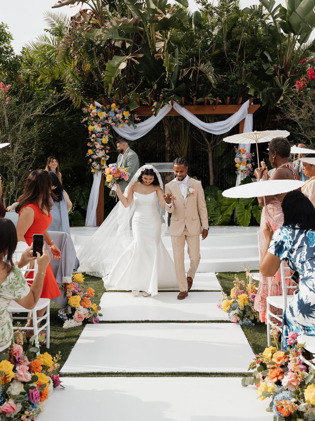 Bride and groom walk down white aisle at outdoor wedding ceremony with colorful floral arrangements.