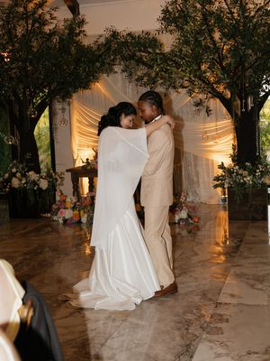 A bride and groom share a romantic first dance under glowing trees with white draping at an elegant wedding venue.