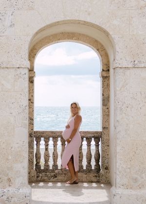 Woman in pink dress poses in series of arched stone balconies overlooking ocean view.