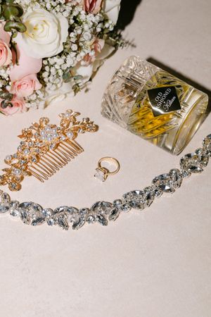 Wedding accessories including delicate jewelry, floral arrangements, perfume bottles and hair combs laid out on white surface.