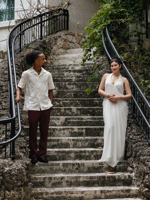 Couple in elegant attire standing on stone steps with wrought iron railings, man in white shirt and woman in white gown.