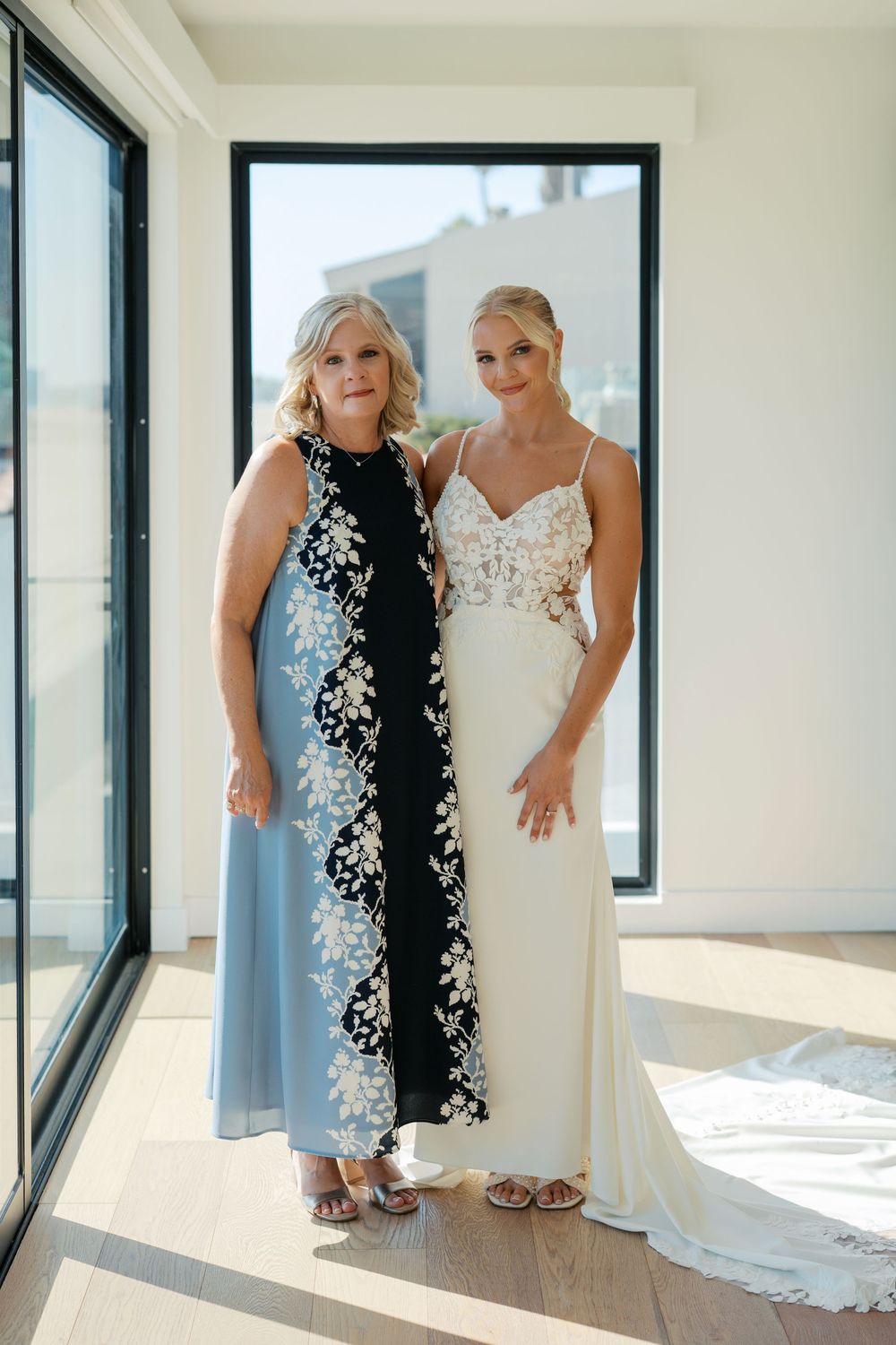 Two people posing together in formal wear, one in a black floral dress and one in a white gown.