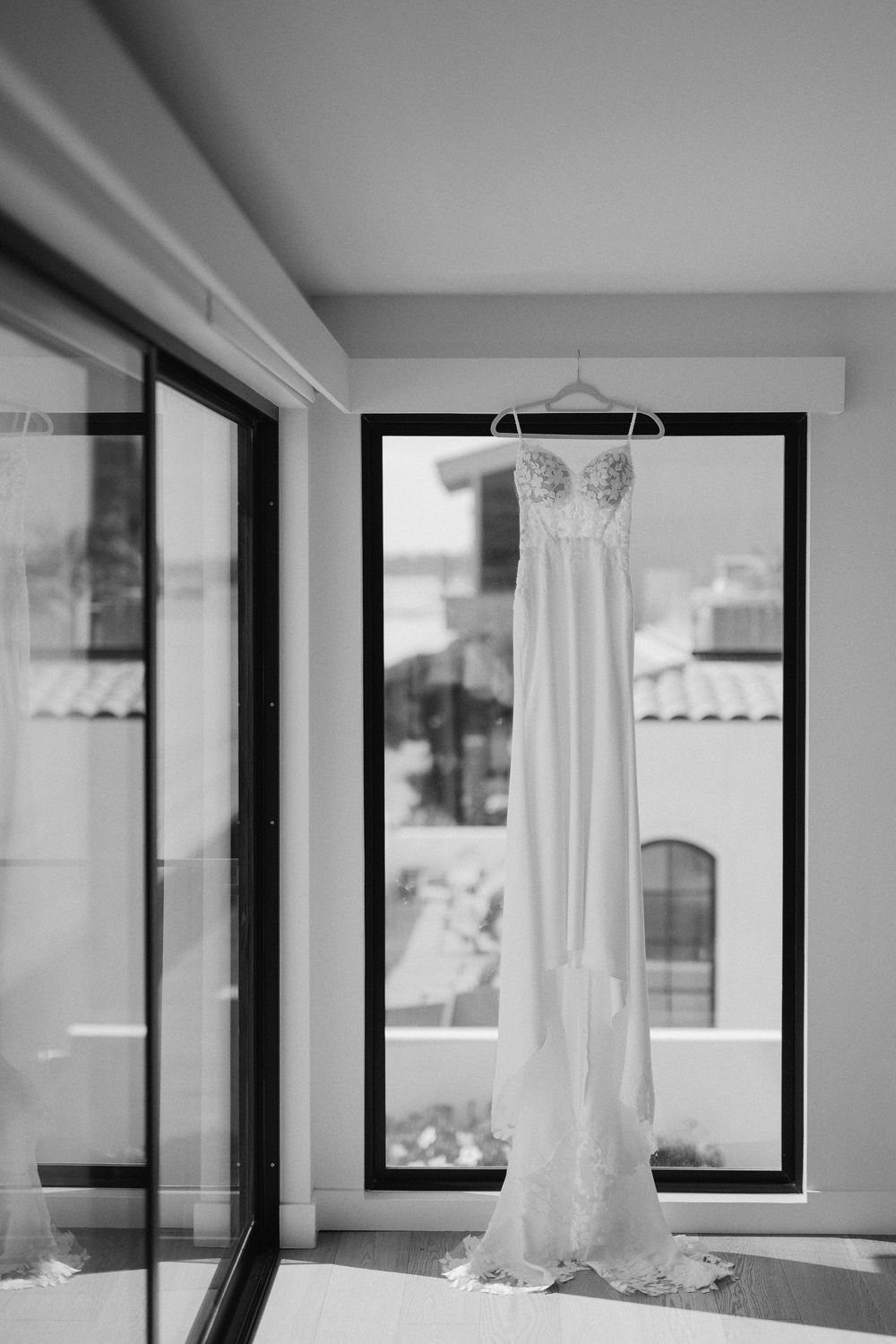 A white wedding dress hangs elegantly near large windows in a bright, modern room with sunlight streaming in.
