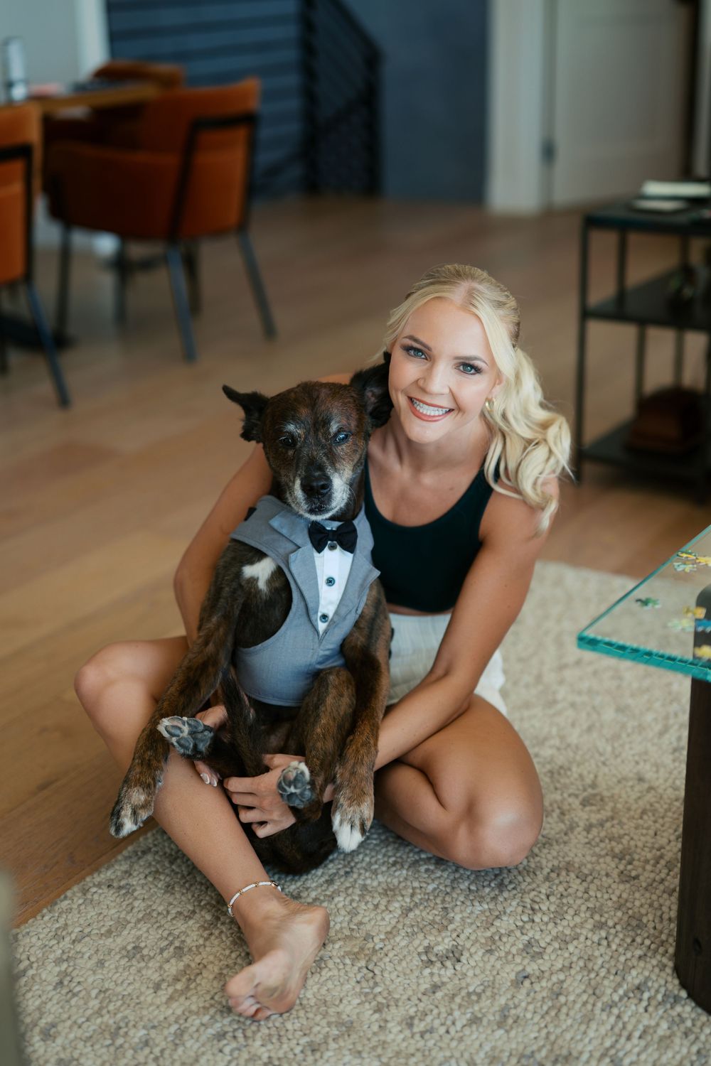 A dog wearing a formal grey tuxedo vest sits with someone on a carpeted floor in a modern living room setting.