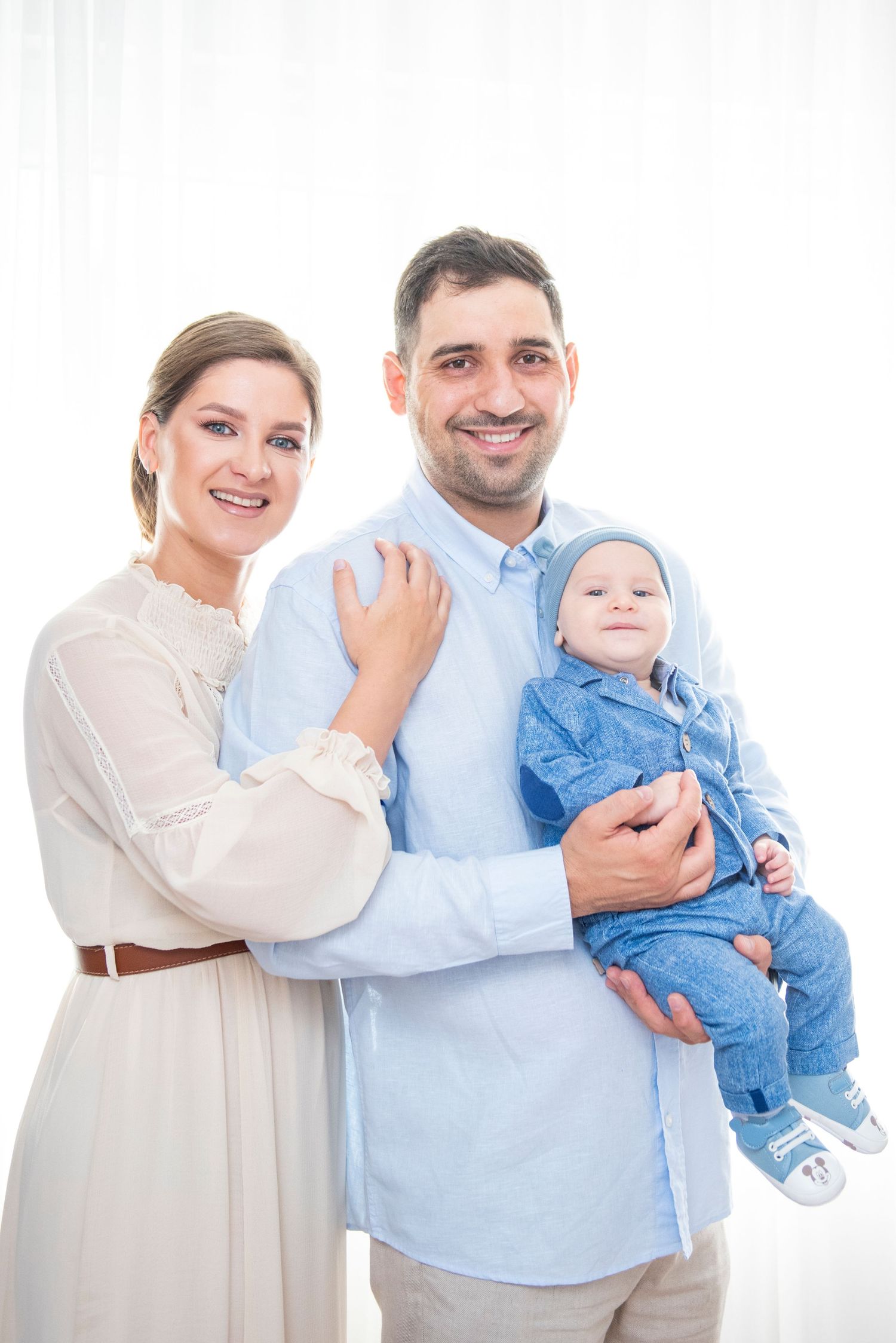 Family portrait with parents in light colors holding baby in blue outfit against white background.