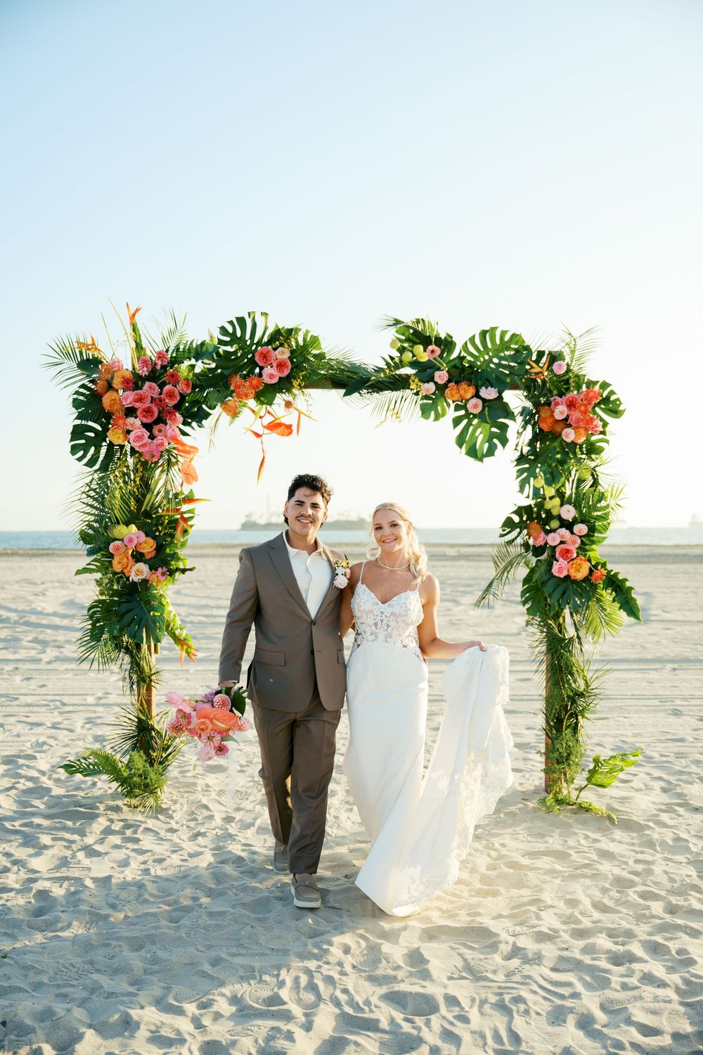 A beach wedding ceremony under a tropical floral arch with monstera leaves and coral flowers on a sandy shore.