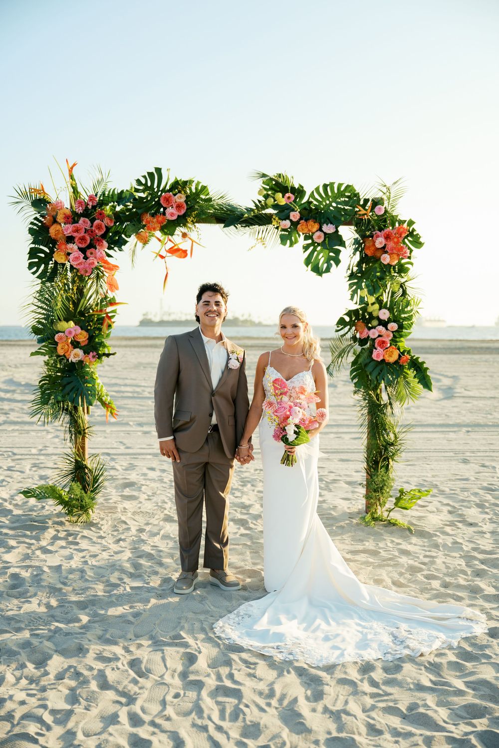 A beach wedding ceremony with a tropical flower arch adorned with pink blooms and greenery on a sandy shore.