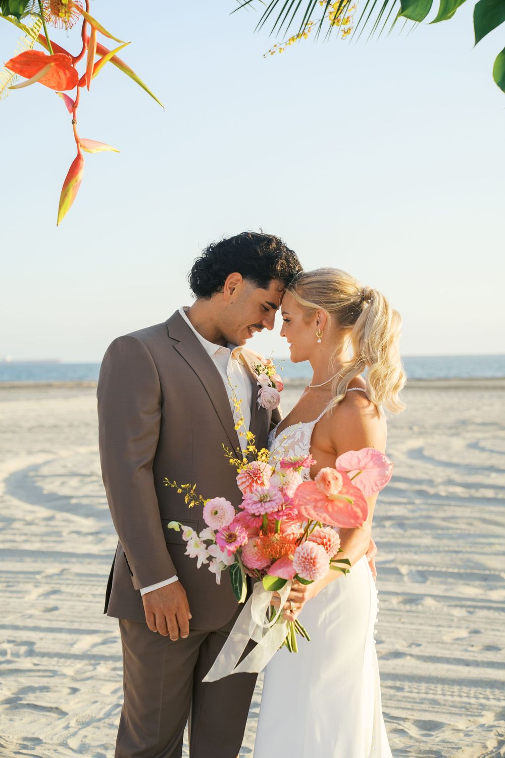 Romantic beach wedding couple embracing at sunset with tropical flowers and palm trees in background.