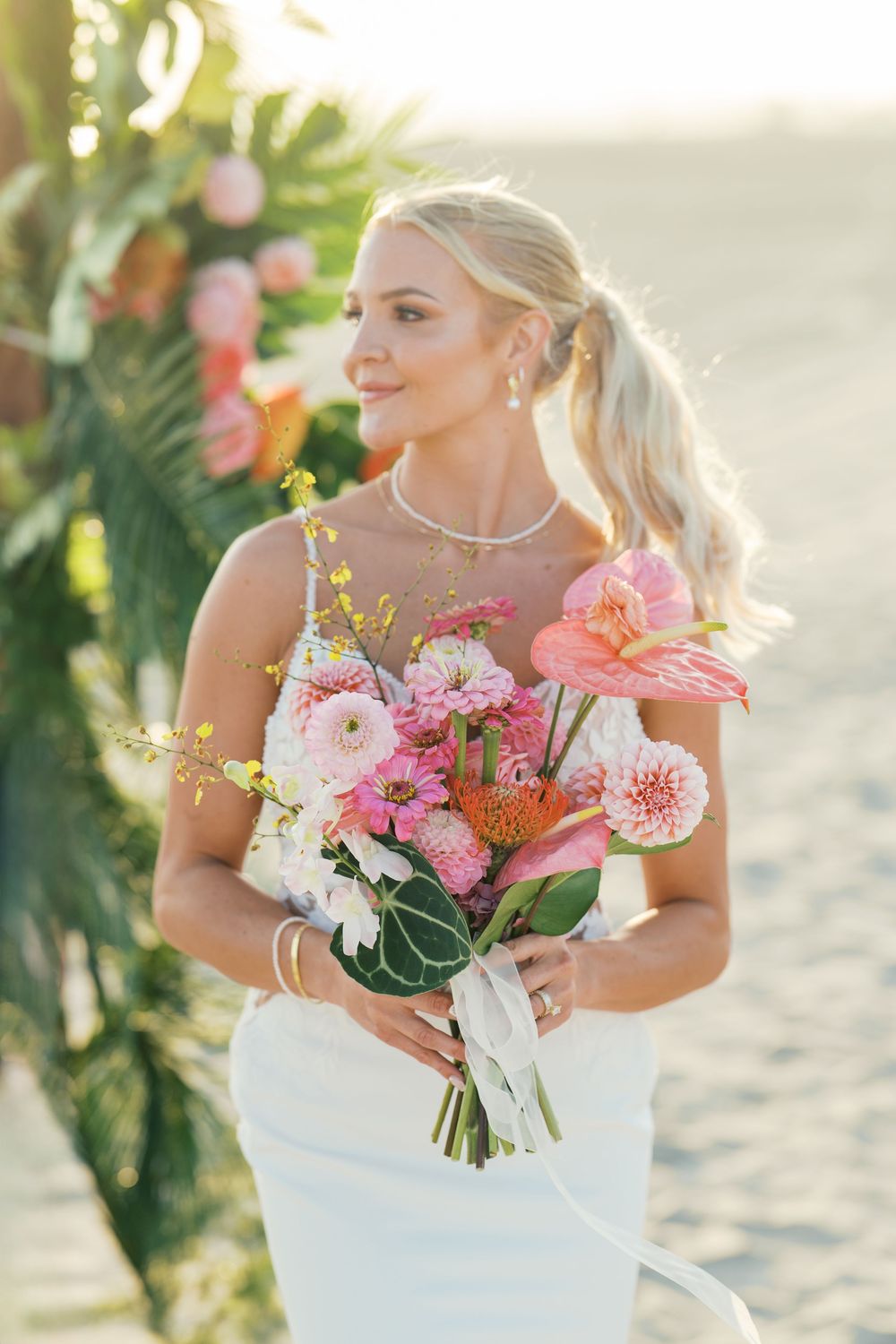 A bride in a white dress holds a vibrant bouquet of pink and coral flowers against a sunlit beach backdrop.