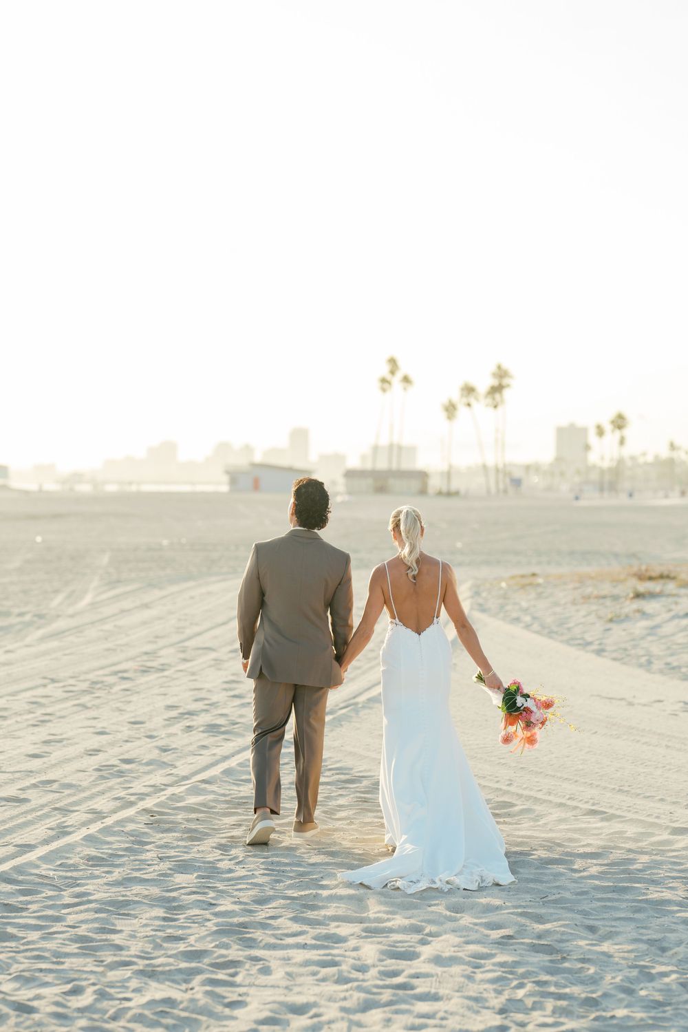 Newlywed couple walking hand in hand along white sandy beach during sunset ceremony.