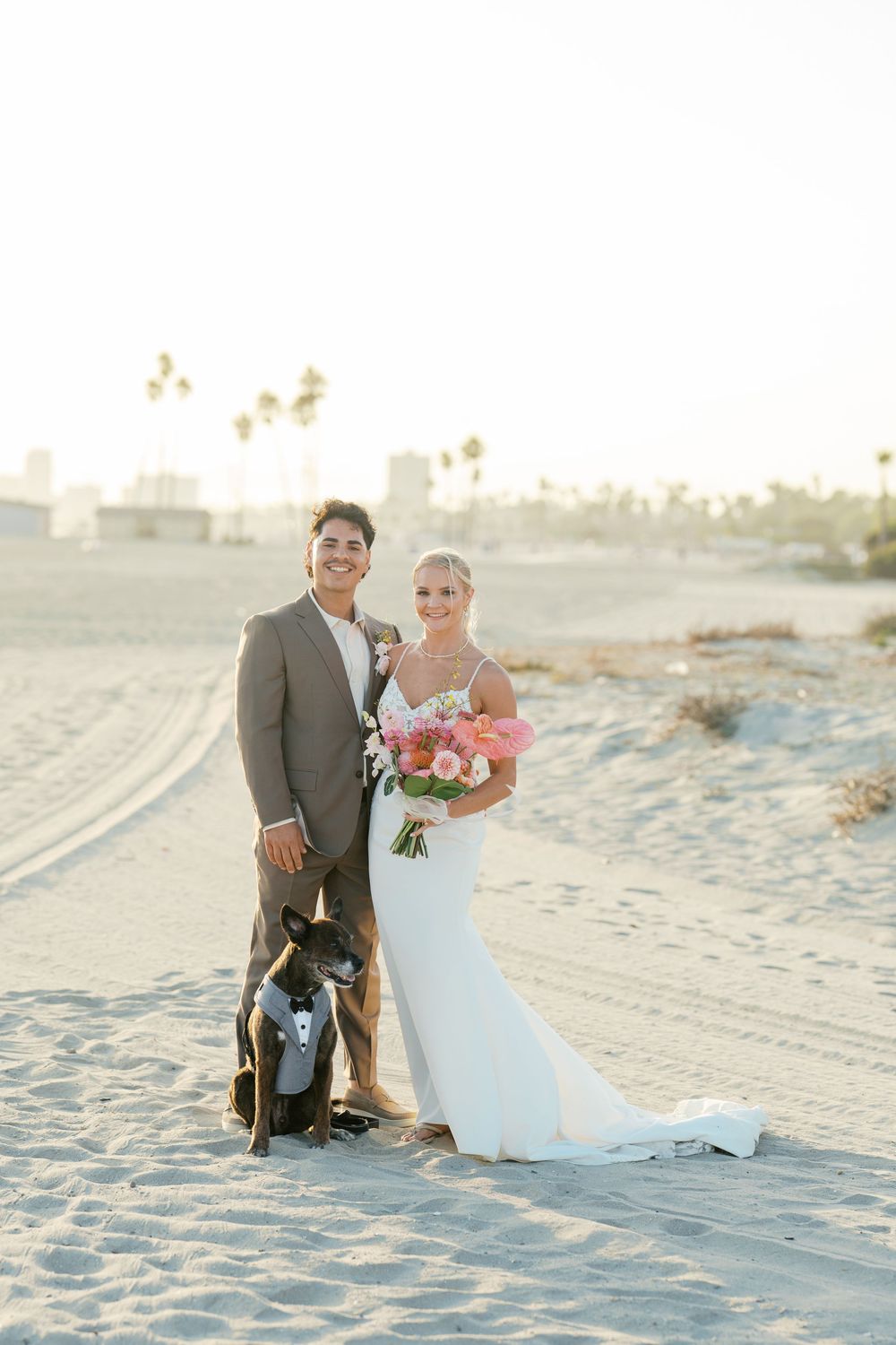 A couple poses with their dog on a sunlit beach during their wedding photo session at sunset.