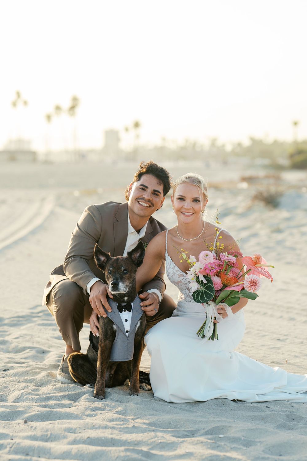 A couple poses with their black dog wearing a bowtie on a sunny beach during their wedding photoshoot.
