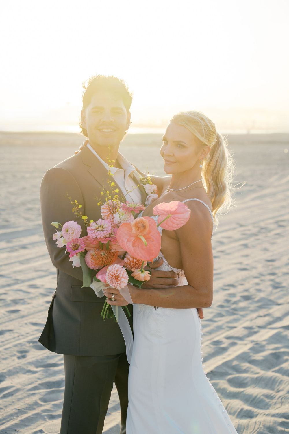 Romantic sunset beach wedding couple embraces while holding a vibrant pink and coral floral bouquet on white sand.