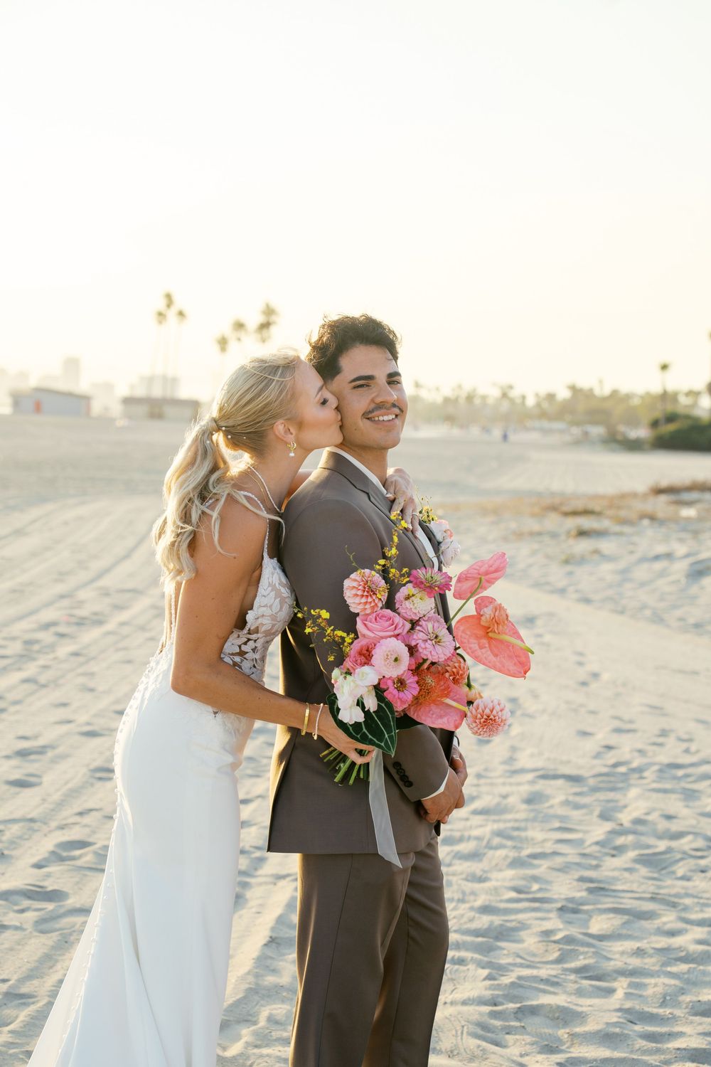 Beach wedding couple shares intimate moments in white dress and grey suit with coral floral bouquet.
